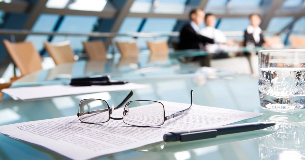 A business meeting in the background with a focus on a legal document and glasses, representing the critical need for a shareholders agreement in business.